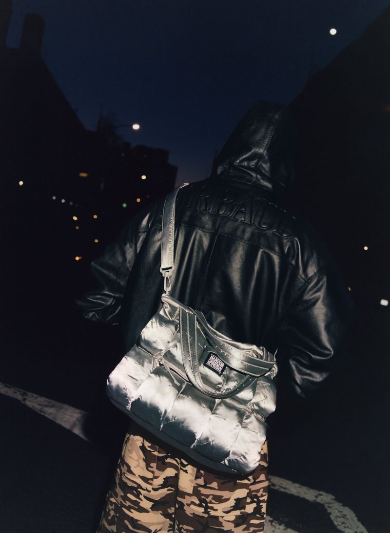 Person with hooded leather jacket and a silver puffy bag crossing a dark city street at night with street lights in background.