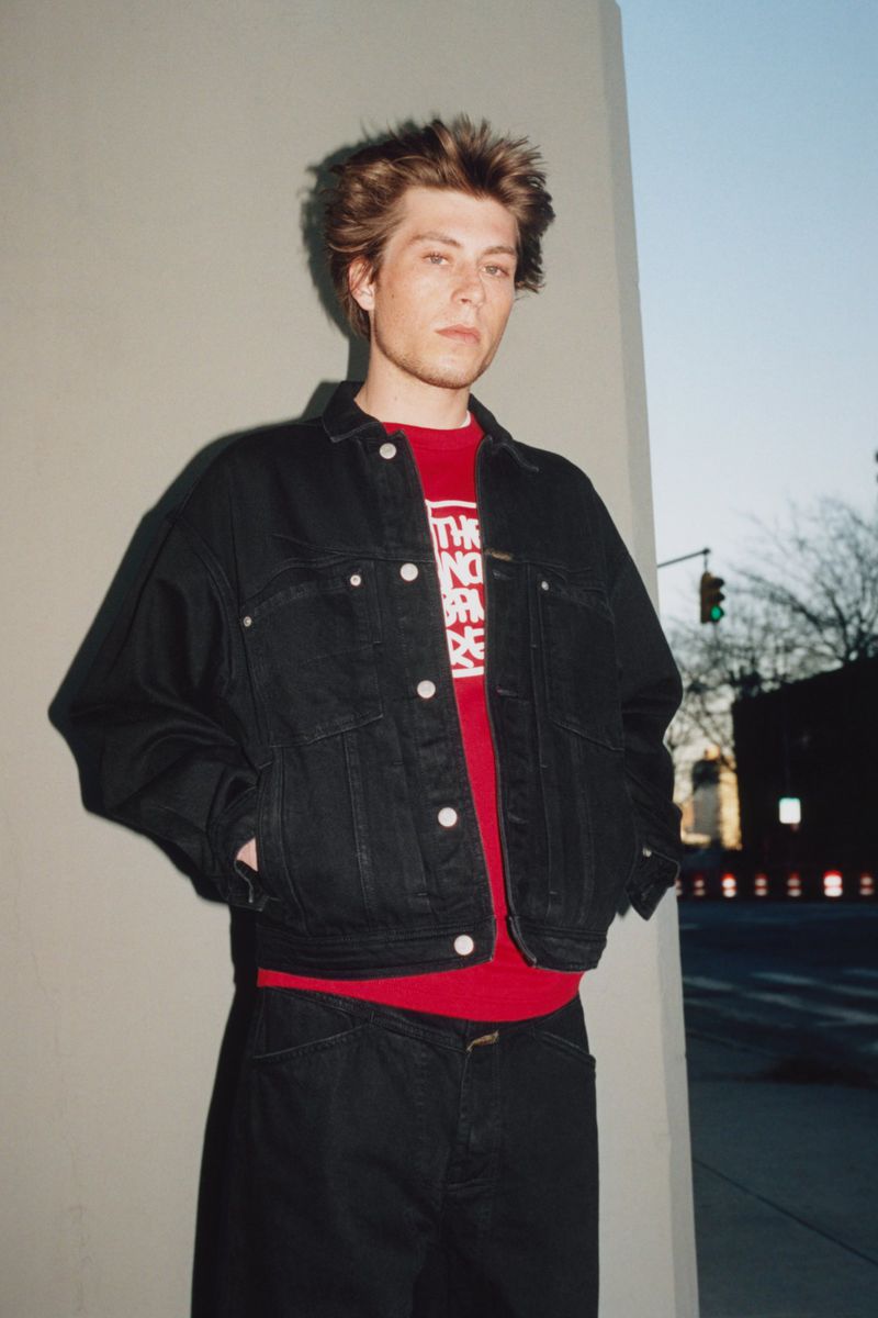 Young adult poses in front of a concrete wall, wearing a denim jacket and a red shirt; cars and buildings are in the background.
