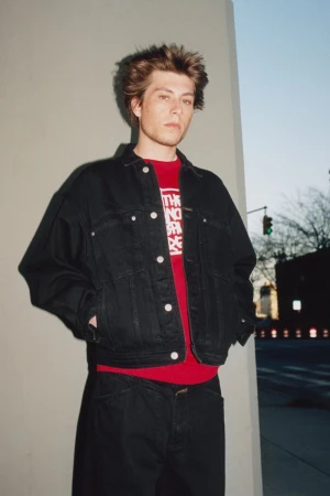 Young adult poses in front of a concrete wall, wearing a denim jacket and a red shirt; cars and buildings are in the background.