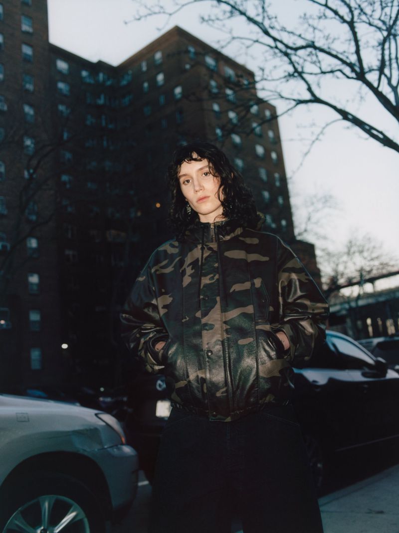 Woman in camouflage jacket poses outdoors against a building and car in urban setting during evening with low light.
