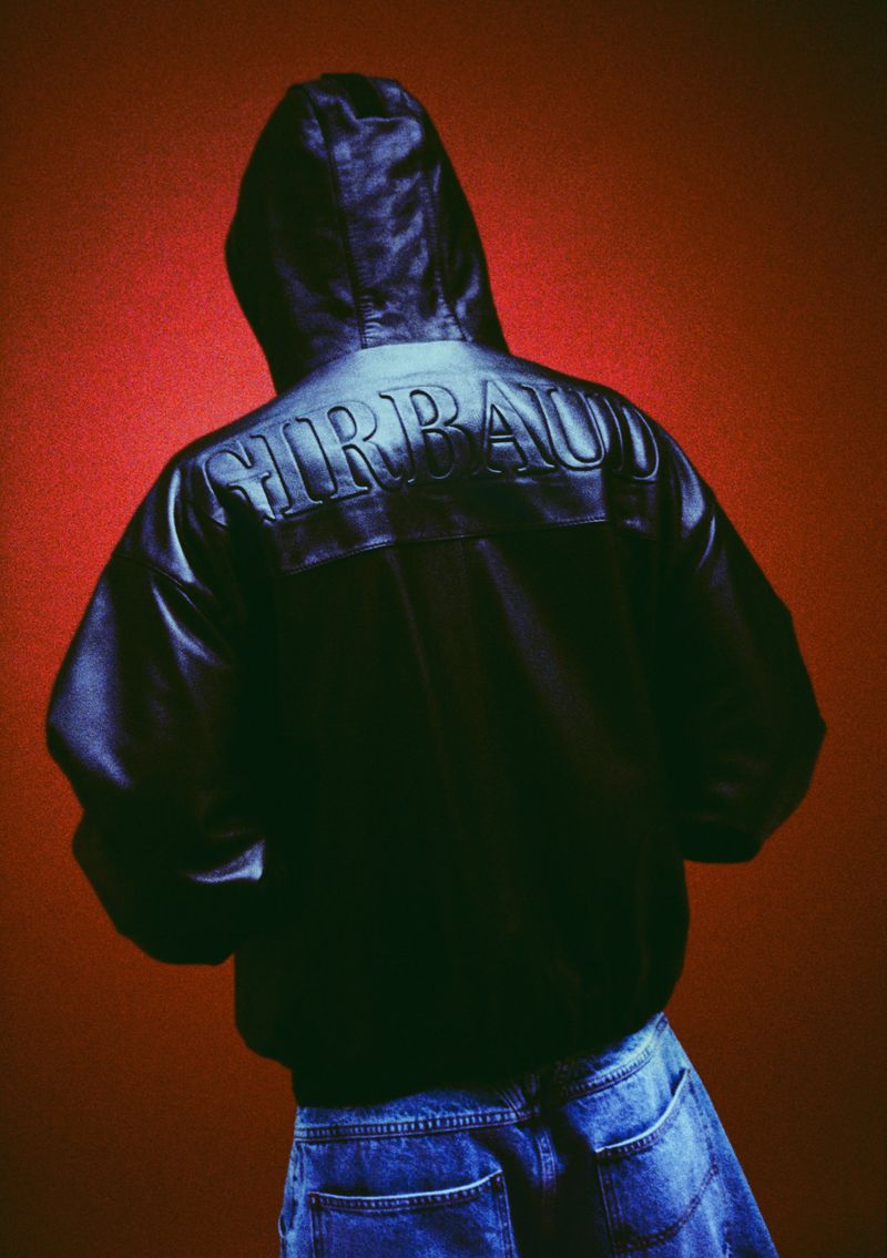 Person wearing a hooded leather jacket and jeans against a red background, showing the jacket's back detail and casual fashion.