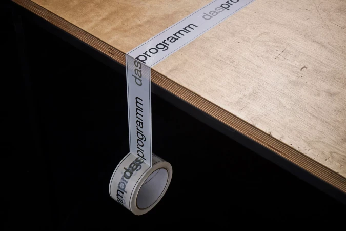 Roll of transparent tape with 'das programm' printed on it is adhered to the edge of a light wood surface against a black background.