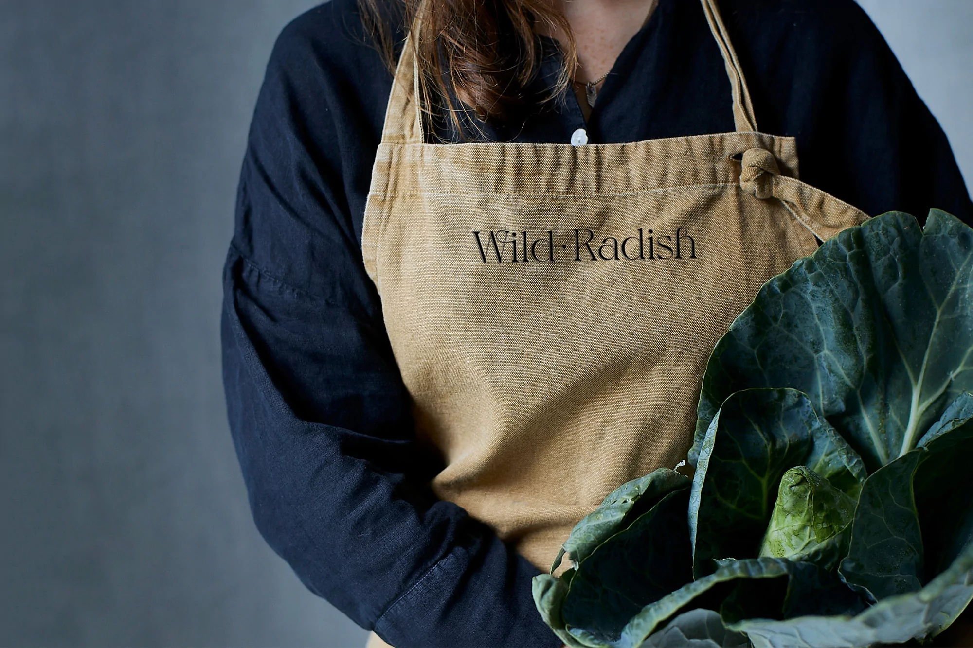 Person in a black shirt and tan apron holds a head of cabbage in a studio setting, featuring the logo “Wild Radish” on the apron.