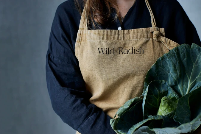 Person in a black shirt and tan apron holds a head of cabbage in a studio setting, featuring the logo “Wild Radish” on the apron.