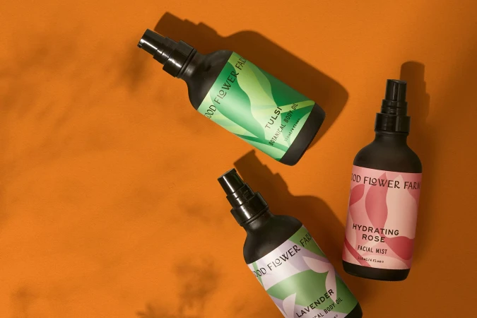 Three matte black spray bottles with botanical labels for Tulsi, Lavender, and Hydrating Rose arranged on a solid orange surface.