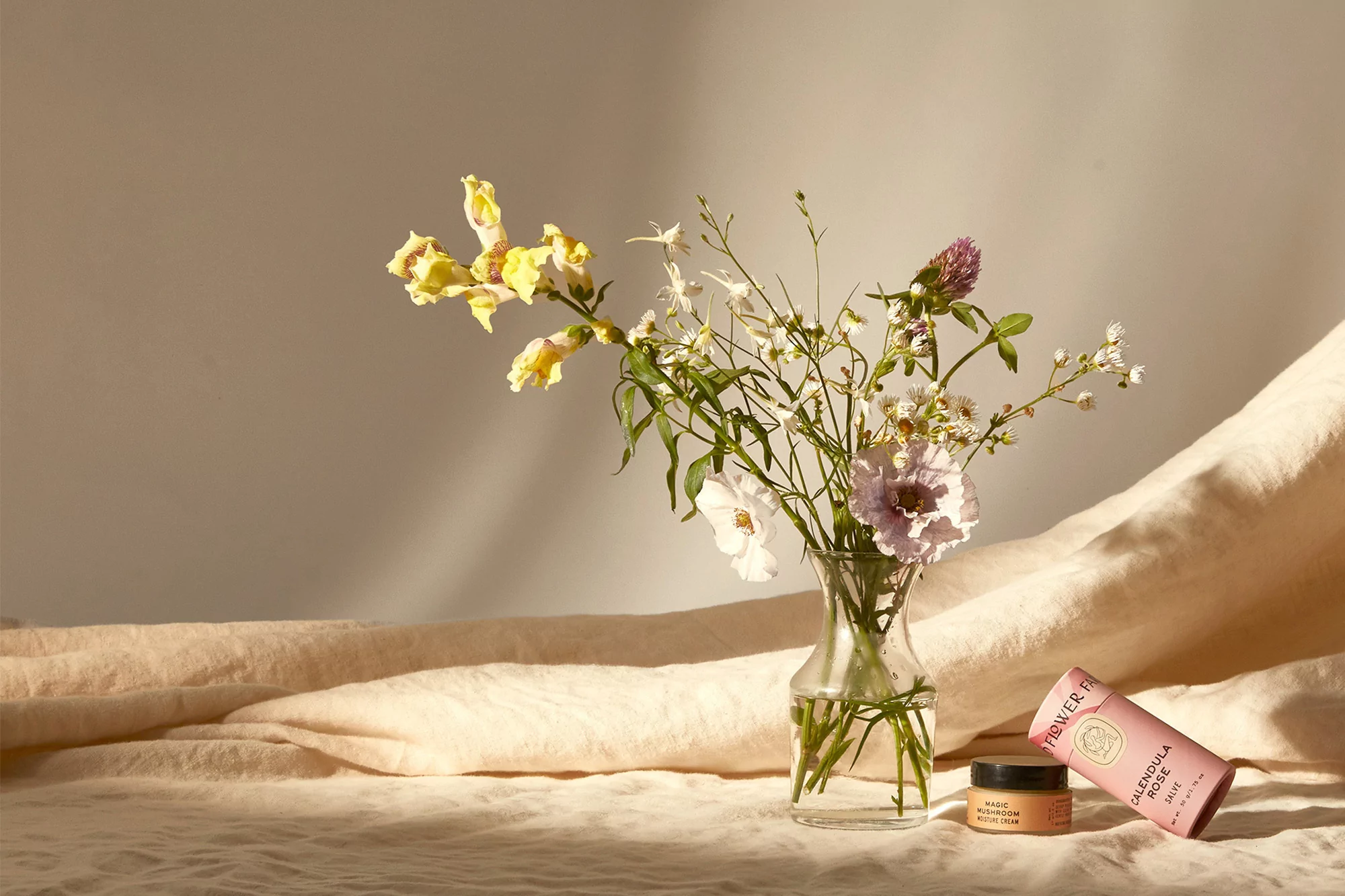Still life of wildflowers in a clear vase with cosmetic product tubes and jars on a draped fabric surface, soft lighting.