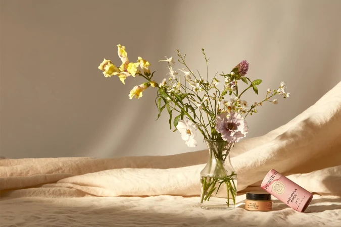 Still life of wildflowers in a clear vase with cosmetic product tubes and jars on a draped fabric surface, soft lighting.