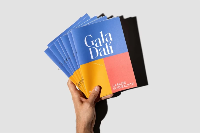Hand holding multiple blue and orange color block magazines titled 'Gala Dalí' against a neutral gray background with hard shadow.