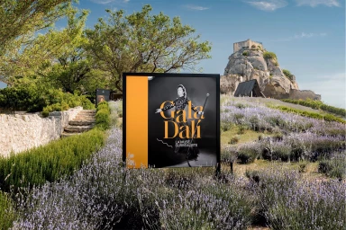 Exhibition sign for Gala Dali La Muse Surrealiste displayed outdoors amidst lush lavender fields under a clear sky.