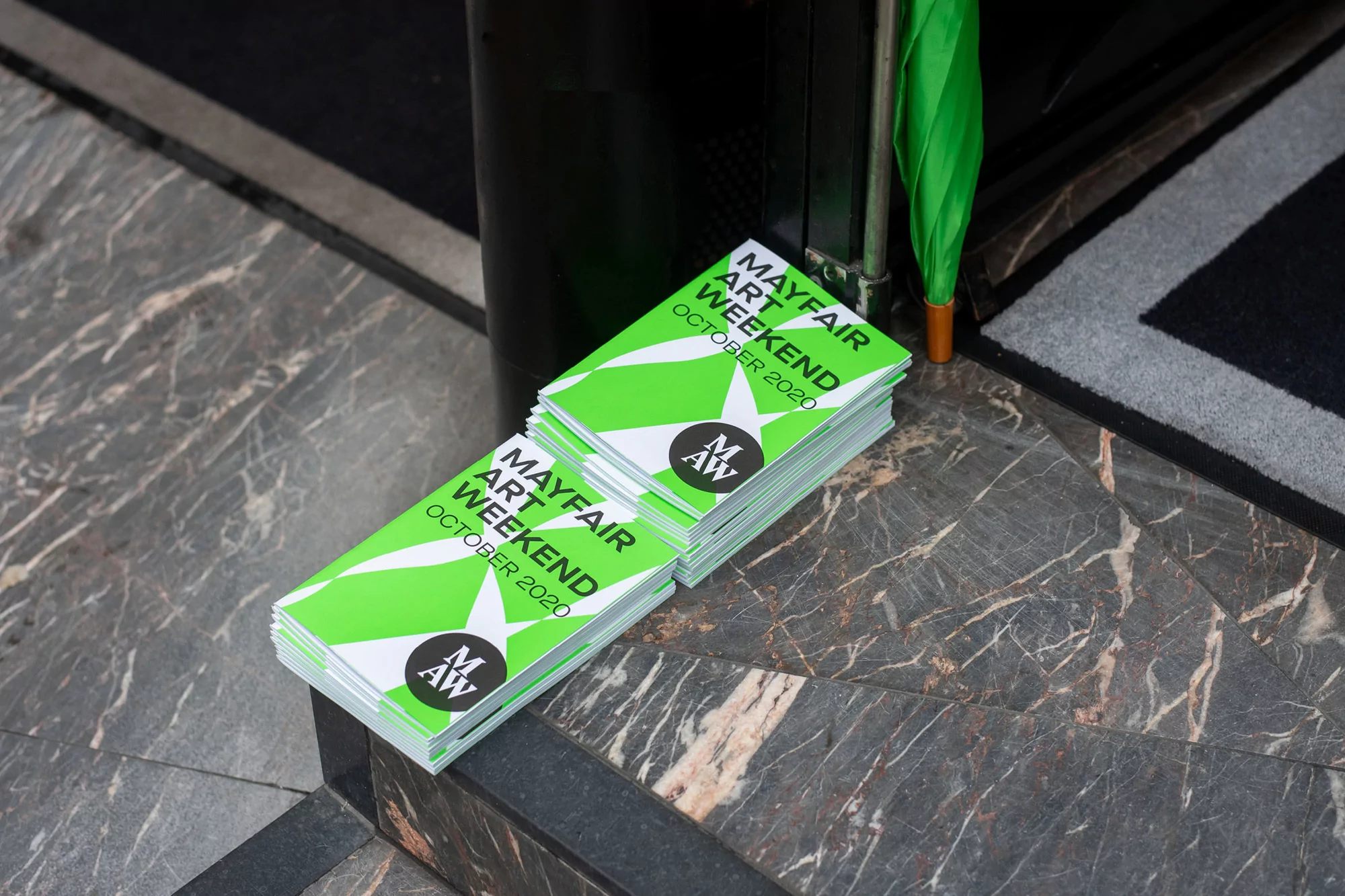 Stacks of Mayfair Art Weekend October 2020 pamphlets on a marble floor next to a black column and green umbrella.