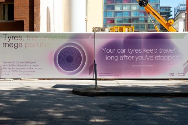 Large outdoor banner advertisement about tyre pollution with purple graphic design mounted on a city street.