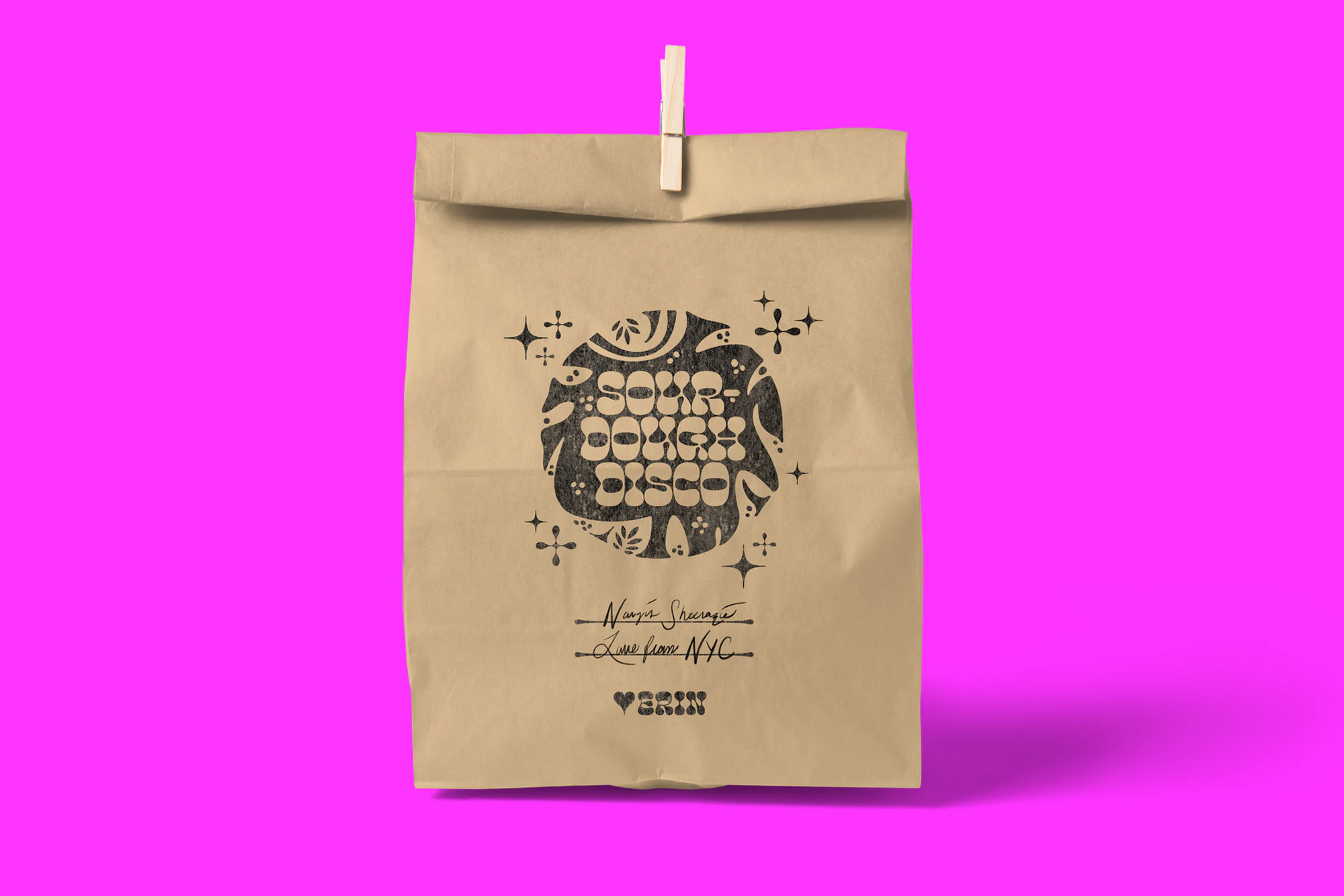 Paper bag with 'Sour Dough Disco' design against a bright pink backdrop, secured with a clothespin. Studio shot with sharp shadows.