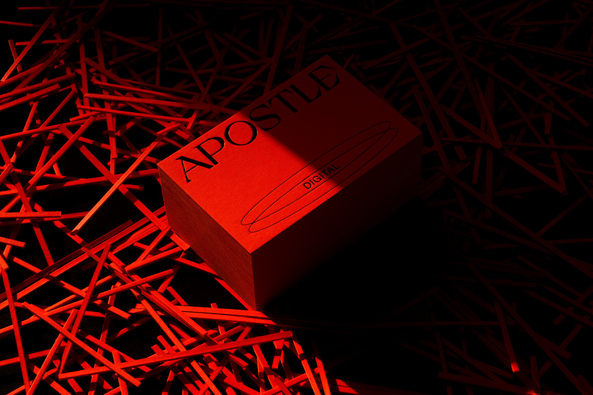 A vibrant red Apostle Digital book sits among scattered red sticks against a dark backdrop, illuminated by studio lighting.