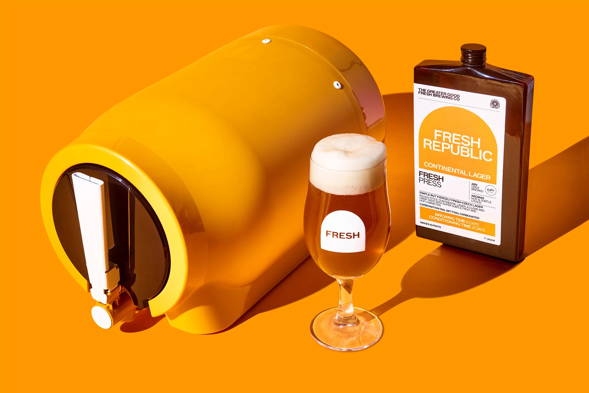 Eye-level product shot featuring a yellow beer dispenser, a glass of frothy beer, and a brown bottle against a bright orange background.
