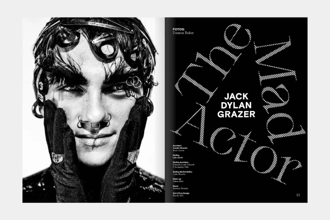 Black and white portrait of a male model with heavy eye makeup, multiple piercings, and hair accessories. Focus on facial details and styling.