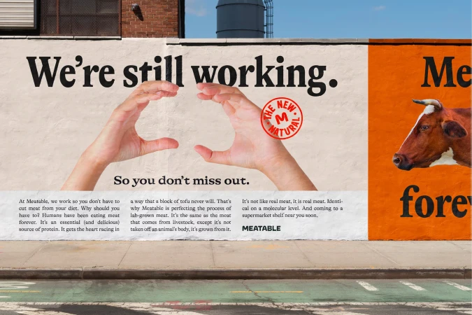 Promotional wall advertisement for Meatable features hands making a heart shape and an image of a cow; the text promotes lab-grown meat.