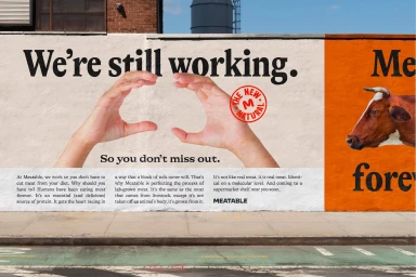Out of home billboard advertisement for Meatable showing two hands framing text and a cow illustration on an exterior brick wall.