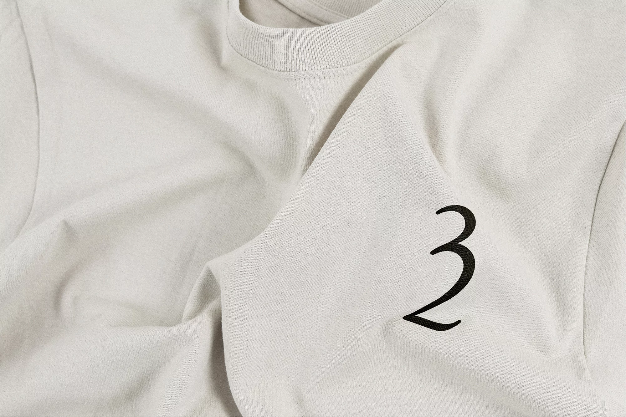 Close-up of a light beige crewneck t-shirt with a large black stylized number '3' on the front, showcasing casual apparel.