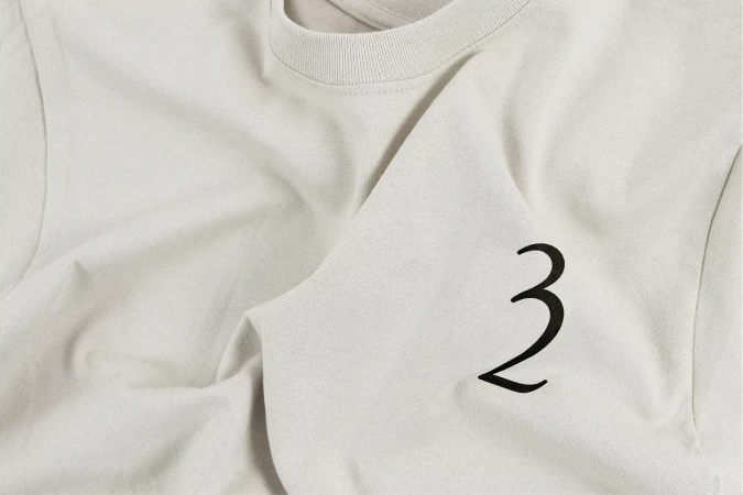 Close-up of a light beige crewneck t-shirt with a large black stylized number '3' on the front, showcasing casual apparel.