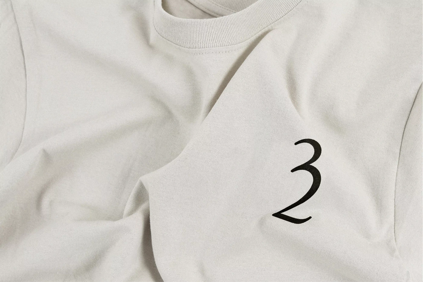 Close-up of a folded off-white cotton t-shirt with a minimalist black numeric logo printed on the fabric.