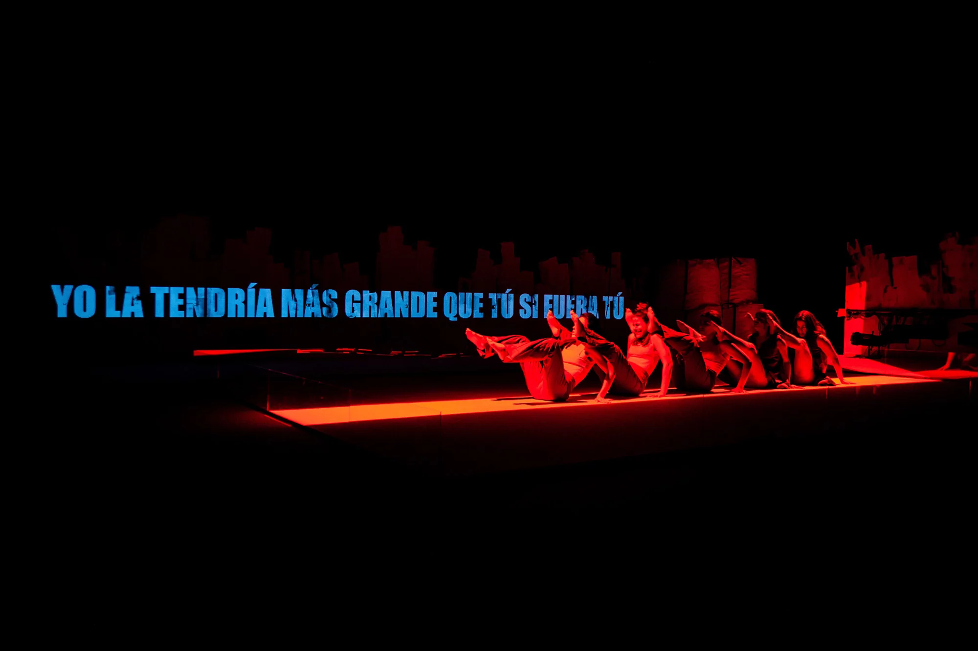 Performers lying on a brightly lit red stage with blue text in Spanish floating against a dark background for Barbara Mestanza brand identity