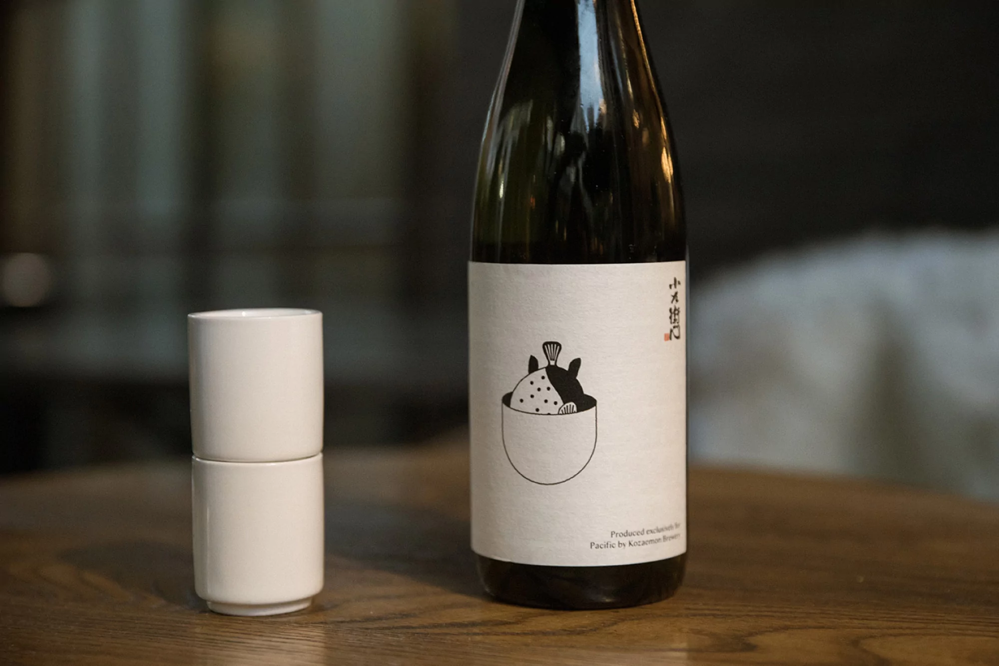 Bottle of sake next to two stacked sake cups on a wooden surface in a softly lit setting. The sake bottle features a minimalist illustration.