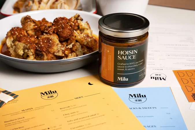 Hoisin sauce jar next to plates and menus featuring Milu restaurant branding, showcasing food and restaurant promotions.