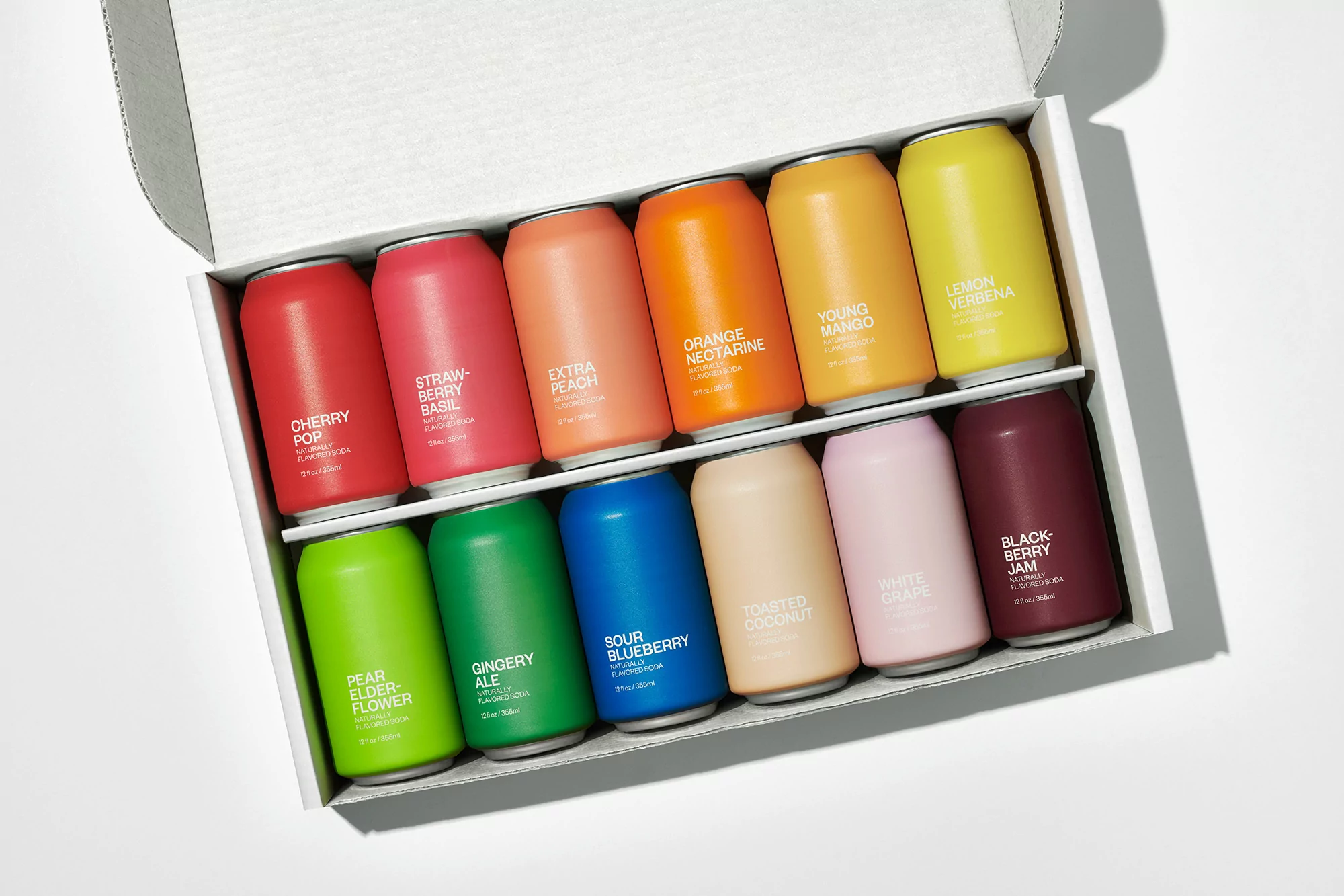 Colorful array of soda cans neatly organized in a box with flavors like cherry, strawberry basil, and lemon verbena on a white background.