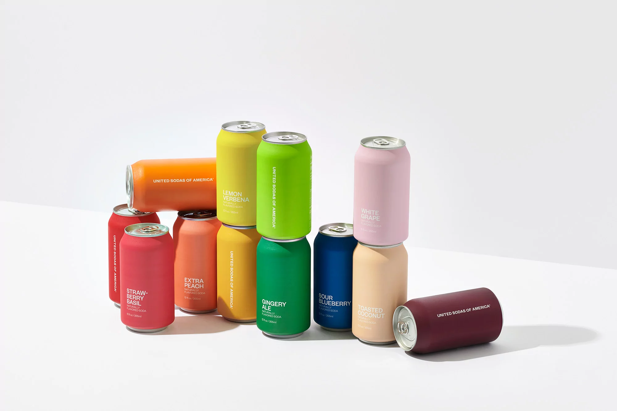 Colorful array of United Sodas of America cans in assorted fruit flavors stacked in a minimalist studio setting with bright, even lighting.
