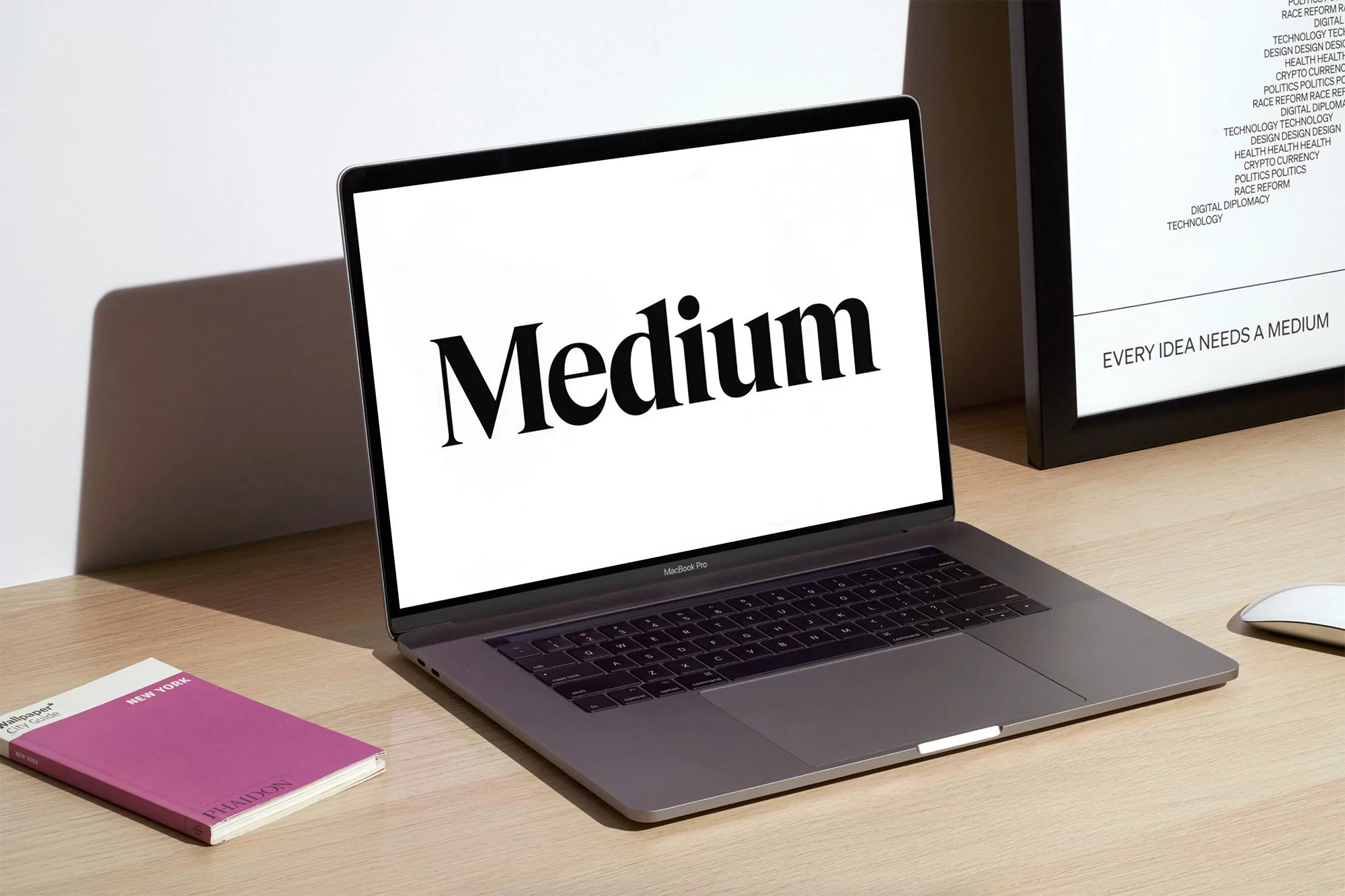 Laptop with 'Medium' displayed on screen, alongside a closed magenta book and a framed poster on a clean, light-toned desk surface.