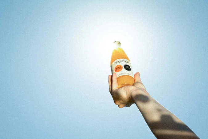 Hand holding a glass bottle of orange juice against a bright blue sky and sun, promoting a fresh, natural beverage outdoors.