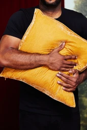 Close-up of a person wearing a black t-shirt holding a large golden yellow velvet throw pillow against their chest.
