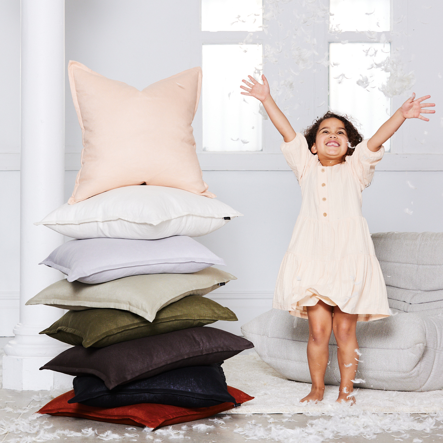 A joyful child plays near a stacked collection of decorative pillows indoors with bright lighting and scattered feathers, creating a playful scene.