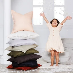 Young child laughing with arms raised beside a tall stack of colorful pillows in a bright studio setting