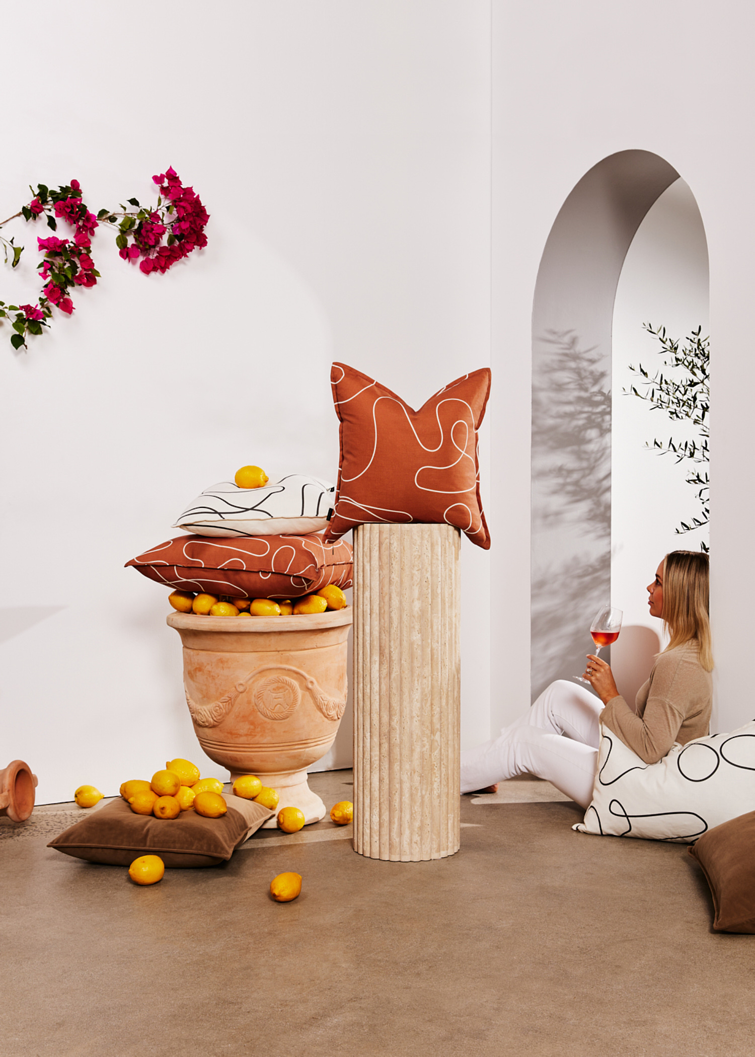 Woman sits near decorative cushions and vase filled with lemons in minimalist setting, holding a glass of rosé wine indoors.