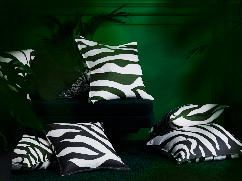 Pile of pillows with a zebra print pattern sits on a dark green couch in front of a dark green wall with tropical plant accents.