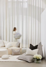 Interior shot featuring a cream sofa, textured cushions, and a motion-blurred person in a white dress near sheer curtains.