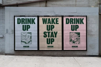 Three posters on a grey wall reading DRINK UP, WAKE UP STAY UP, and DRINK UP for Hokum speciality coffee with illustrated panels.
