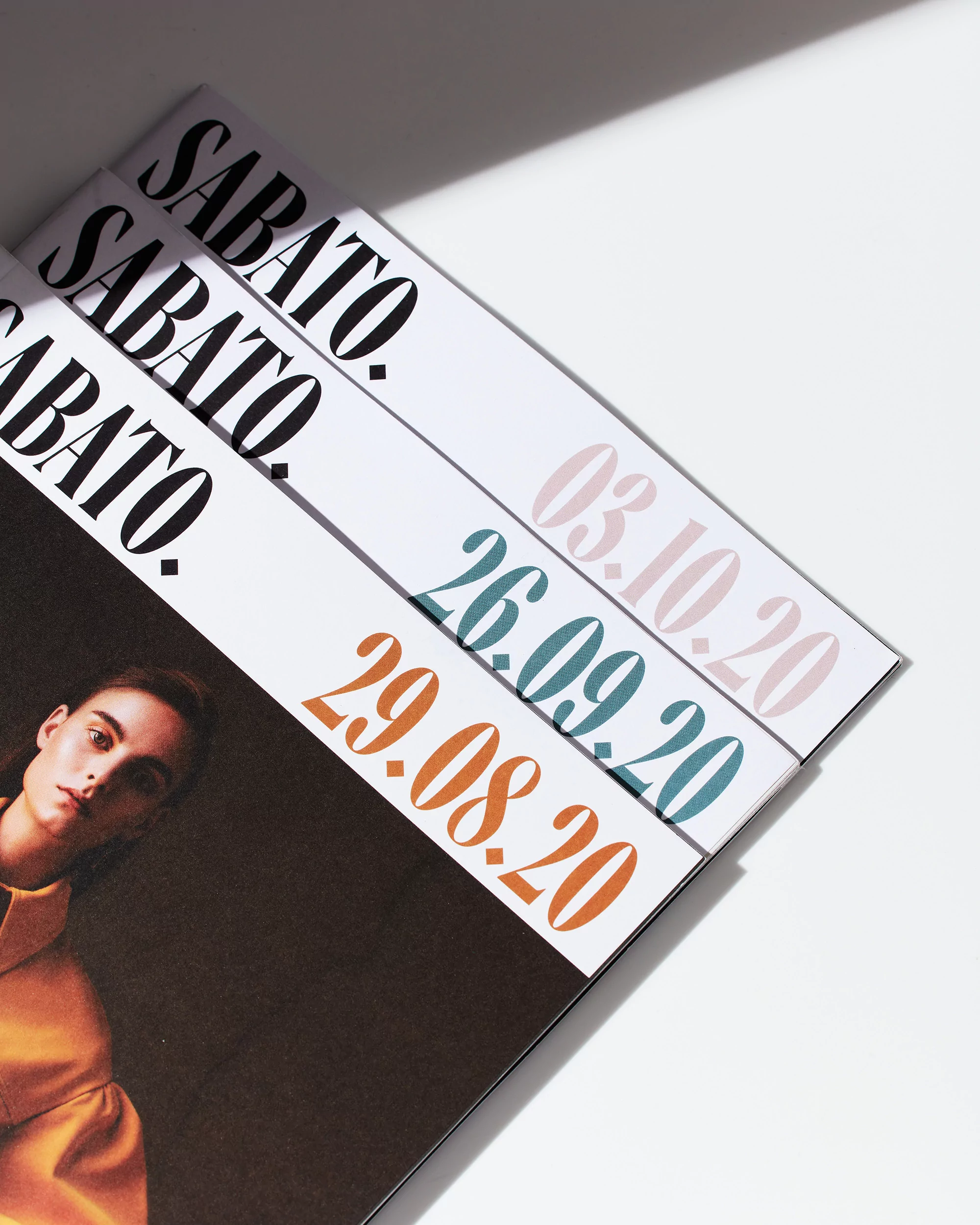 Three white magazines with graphic design lie diagonally in the frame. Magazine cover partially shows a woman with natural makeup.