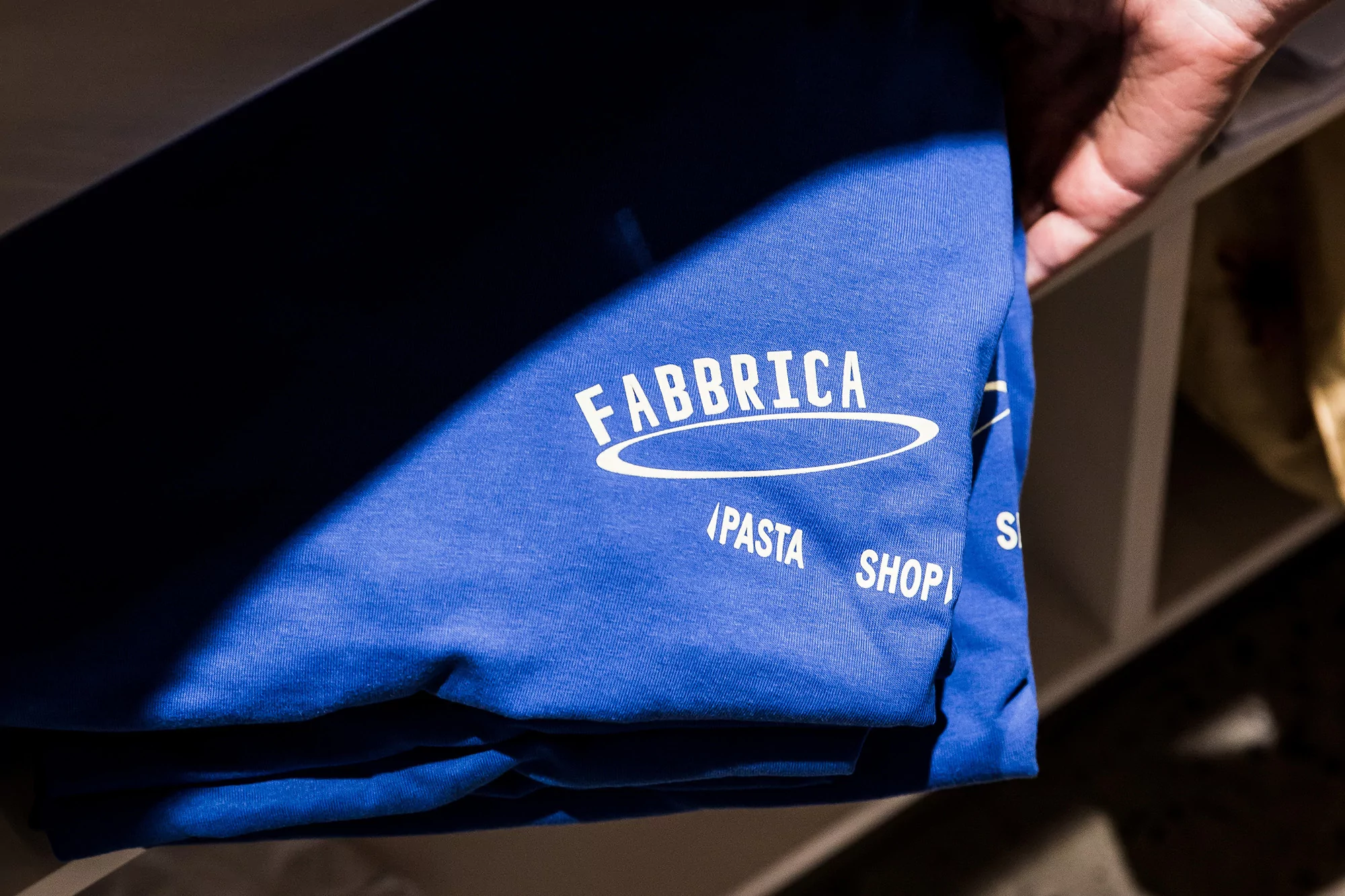 Close-up on blue shirt with 'Fabbrica / Pasta Shop' logo, held by a hand, suggesting a culinary or brand merchandise focus with indoor setting.