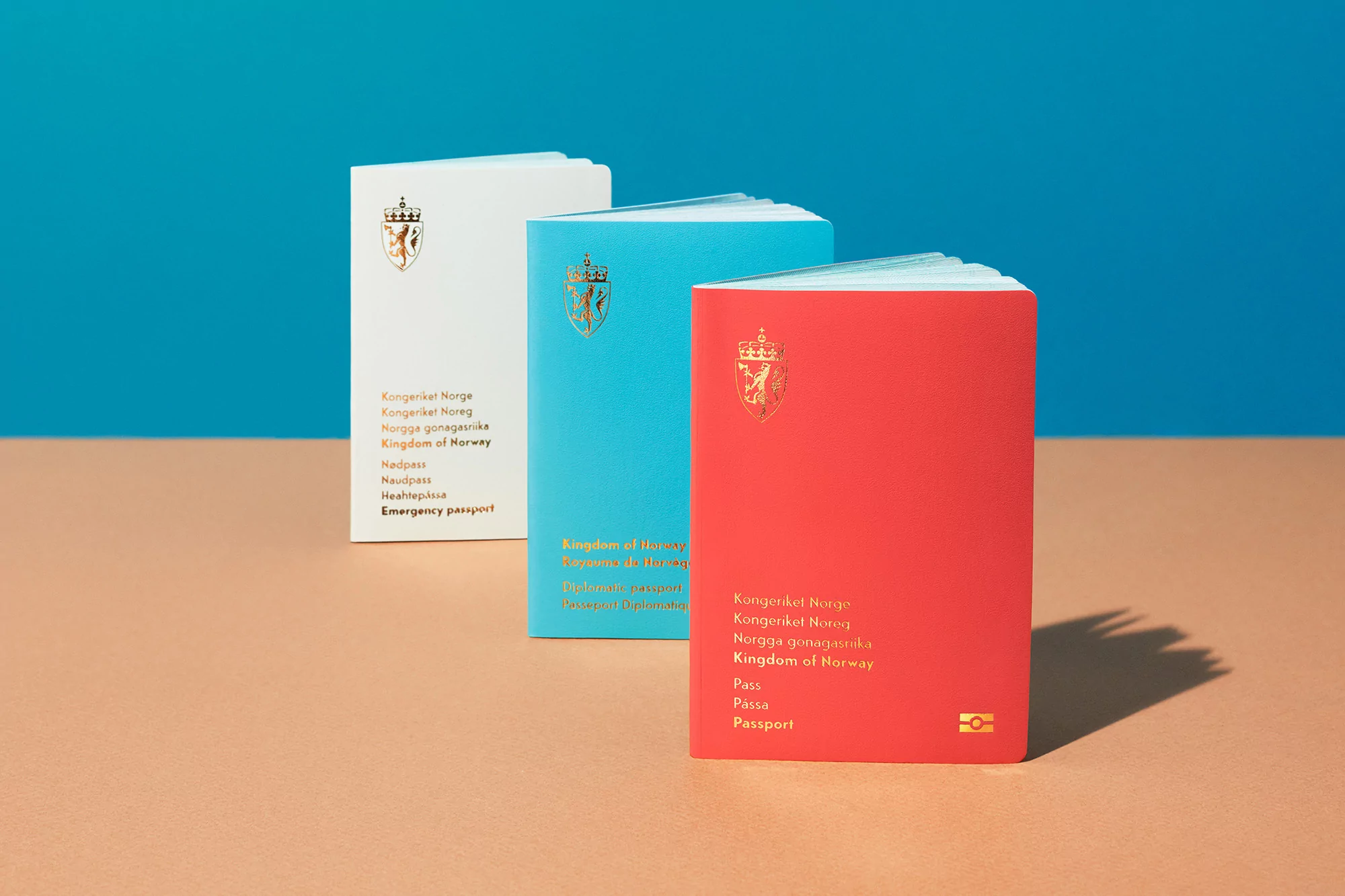Three colorful Norwegian passports (white, blue, and red) are arranged on a tan surface against a blue backdrop, showcasing official travel documents.