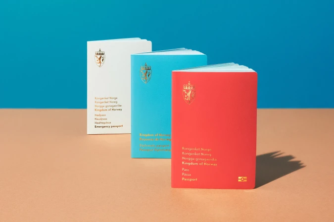 Three colorful Norwegian passports (white, blue, and red) are arranged on a tan surface against a blue backdrop, showcasing official travel documents.
