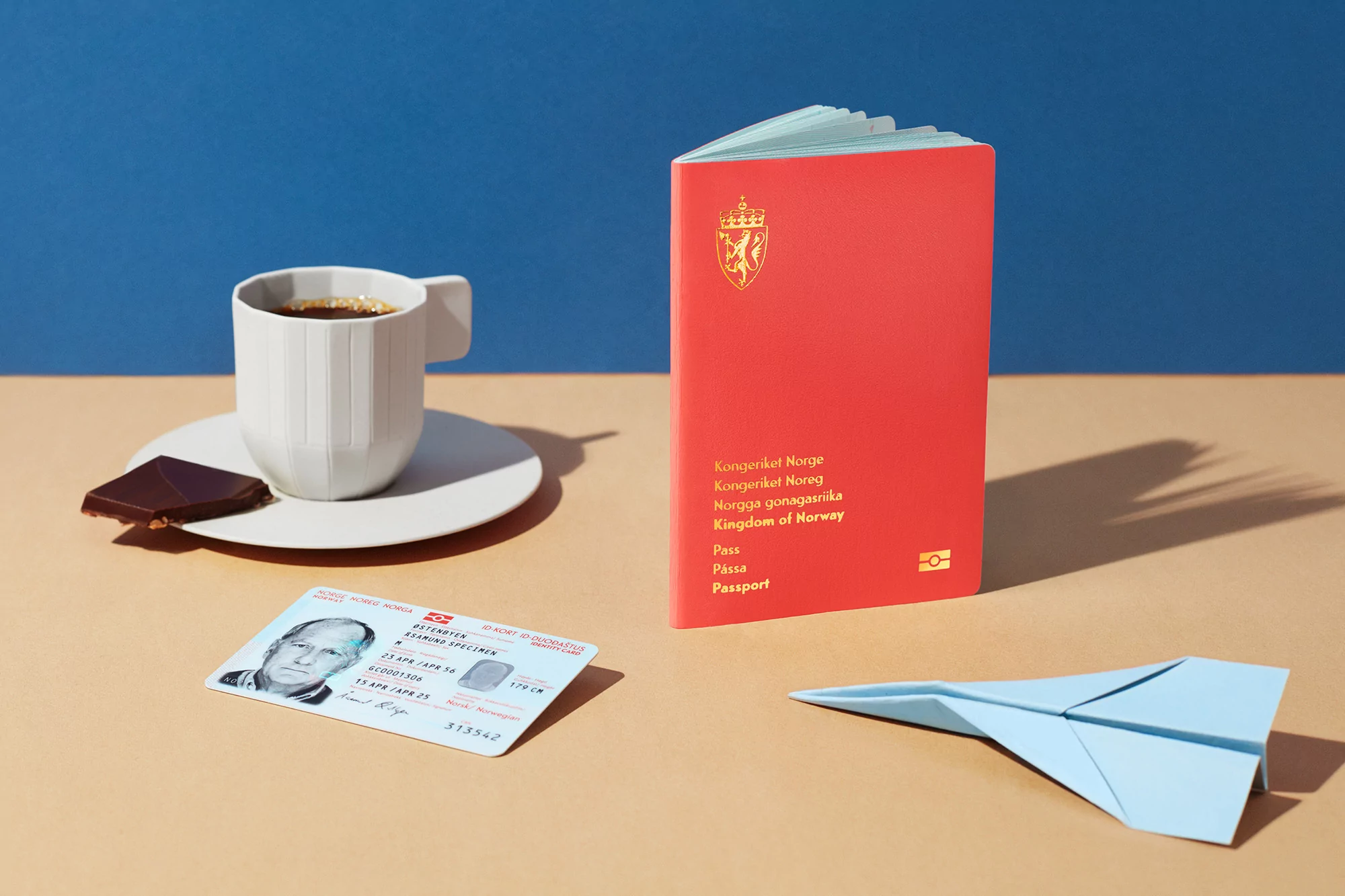 Still life featuring a passport, ID, a cup of coffee, a piece of chocolate, and a paper airplane against a two-tone backdrop.