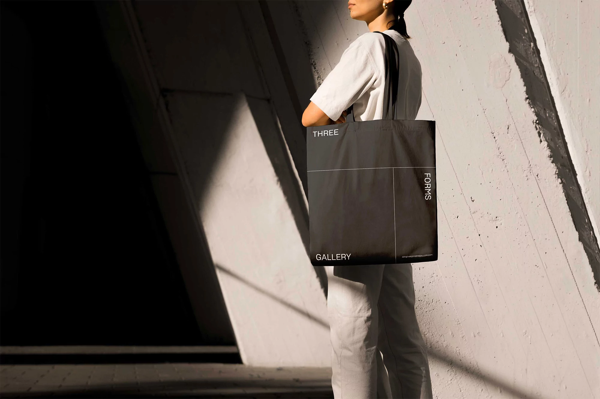 Woman carries black tote bag with white text outdoors against a geometric, light-colored wall with shadows. Minimalist fashion accessory.