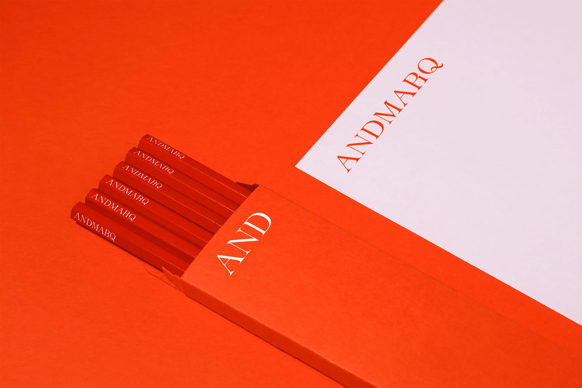 Close-up of red pencils in a box with a white paper card, all featuring the brand name "ANDMARQ", set against a red background.