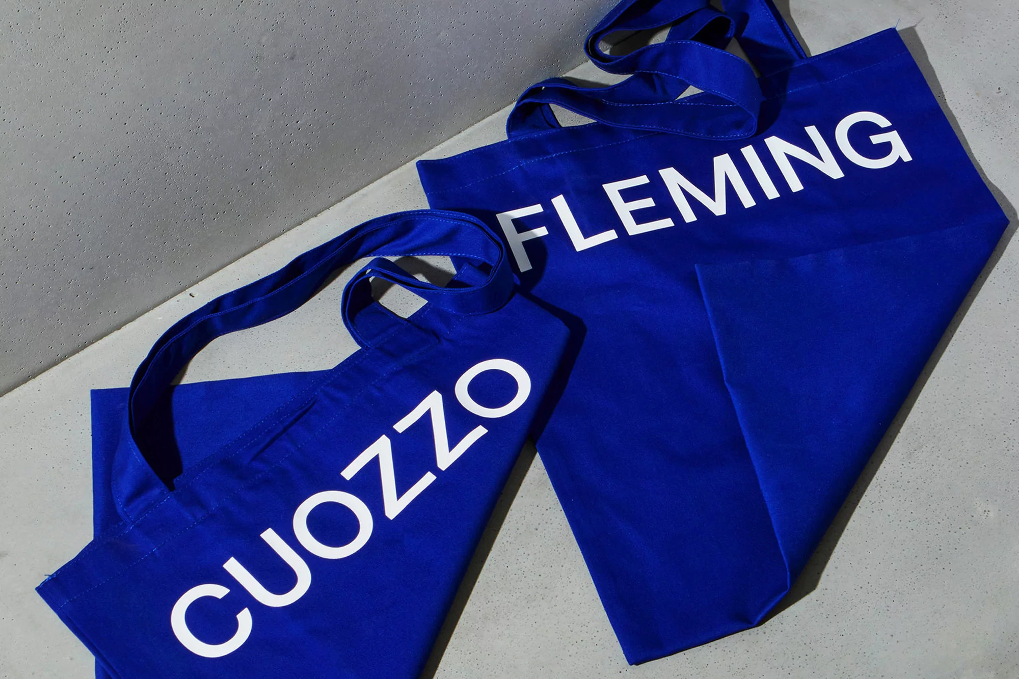 Two blue canvas tote bags, one with 'CUOZZO' and the other with 'FLEMING' in white letters, set against a grey background.