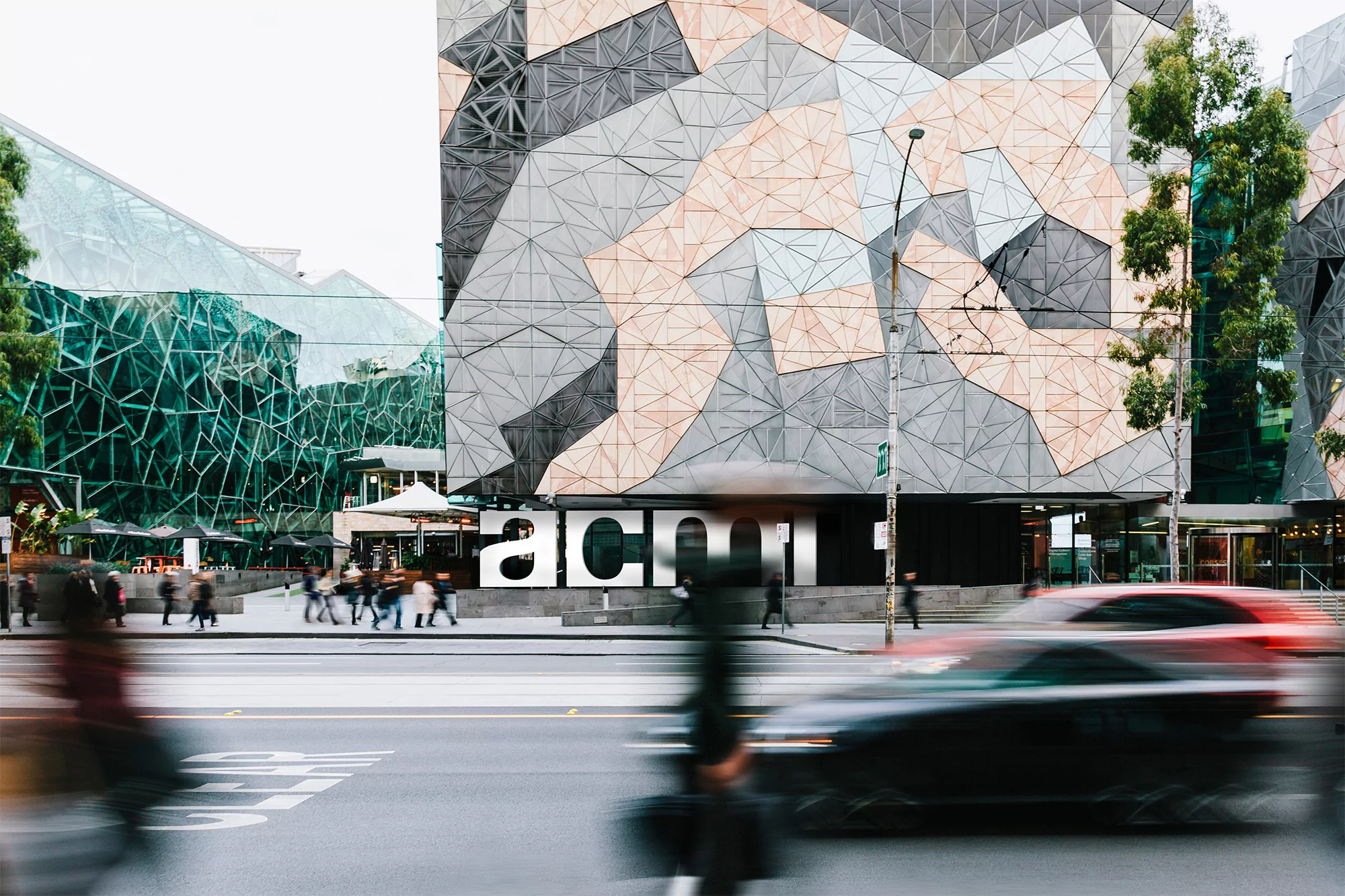 Modern architecture featuring a geometric building facade, pedestrian traffic, and blurred motion of passing vehicles on a city street.