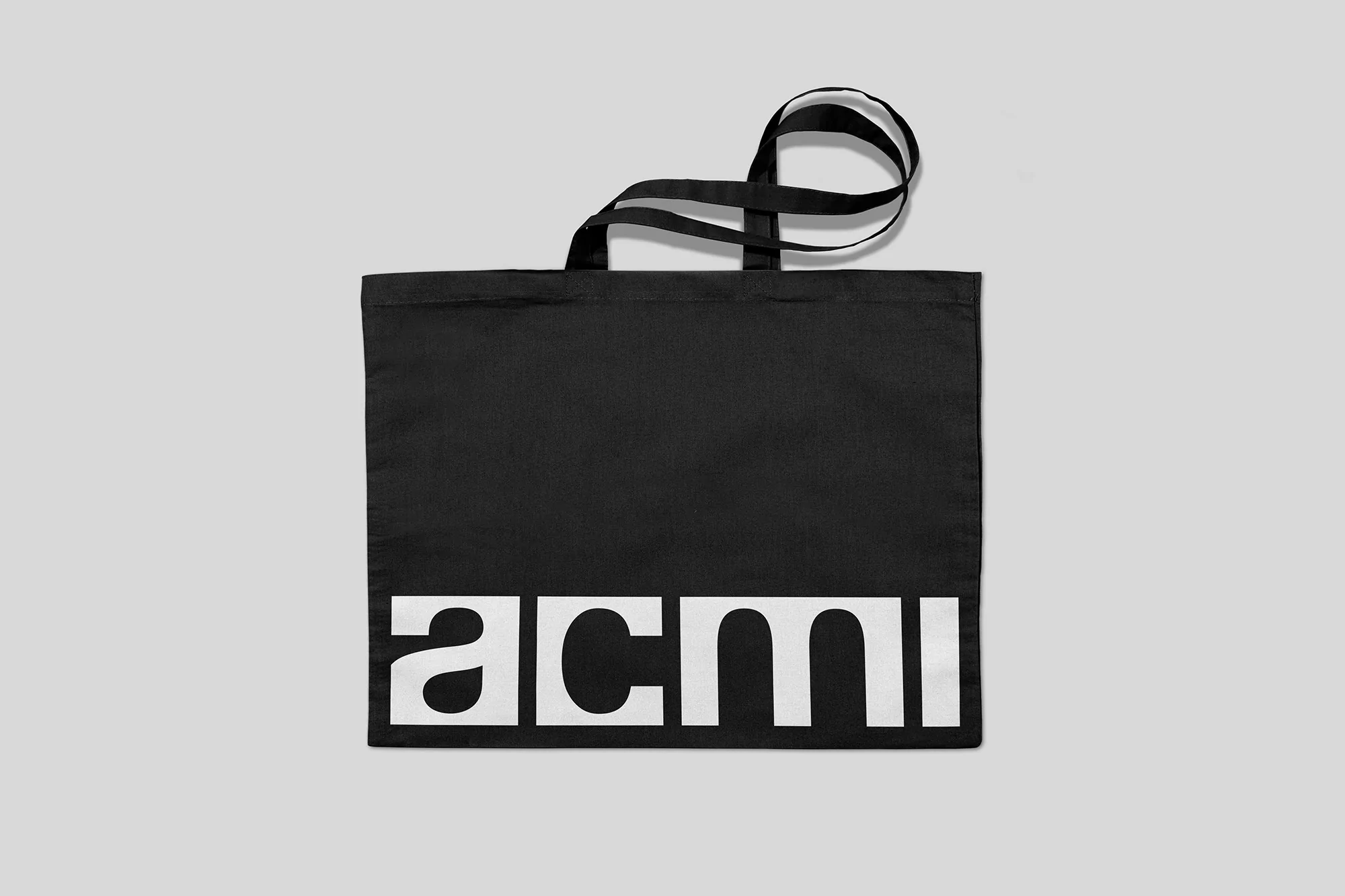 Black tote bag featuring a bold white 'acmi' logo on a light gray background, suitable for shopping or carrying items.