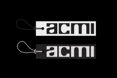 Two rectangular paper price tags featuring bold stencil typography with the word acmi set against a solid black background.