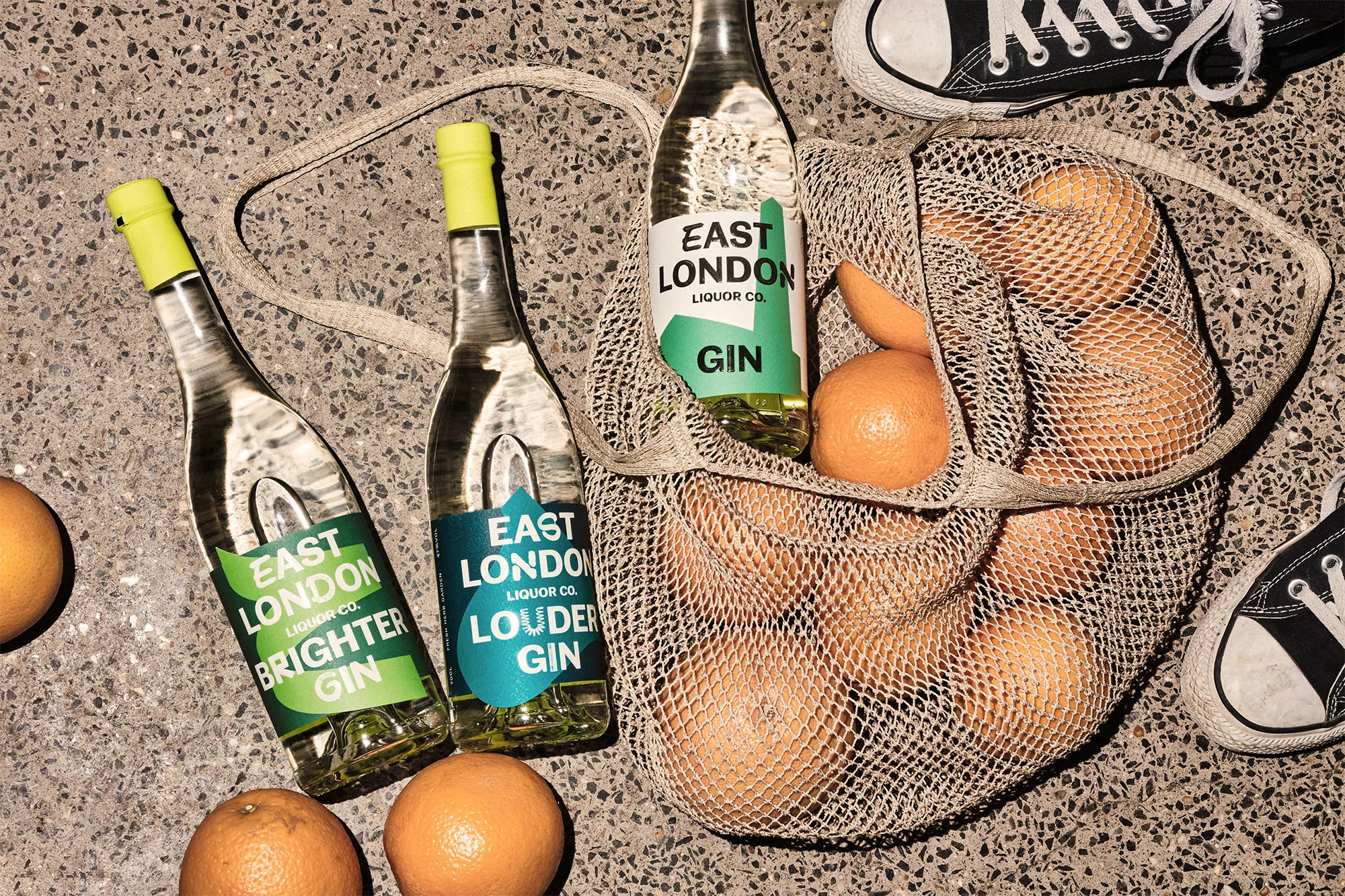 Overhead shot of three bottles of East London gin with oranges in a net bag and black and white sneakers on a speckled surface.
